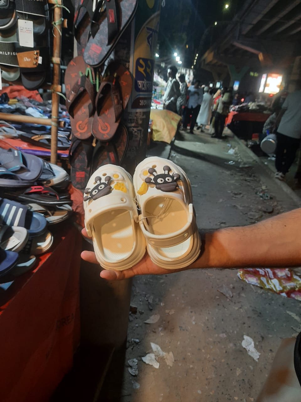 Cream kids clog-style shoes with a large cartoon charm held up at a night street market