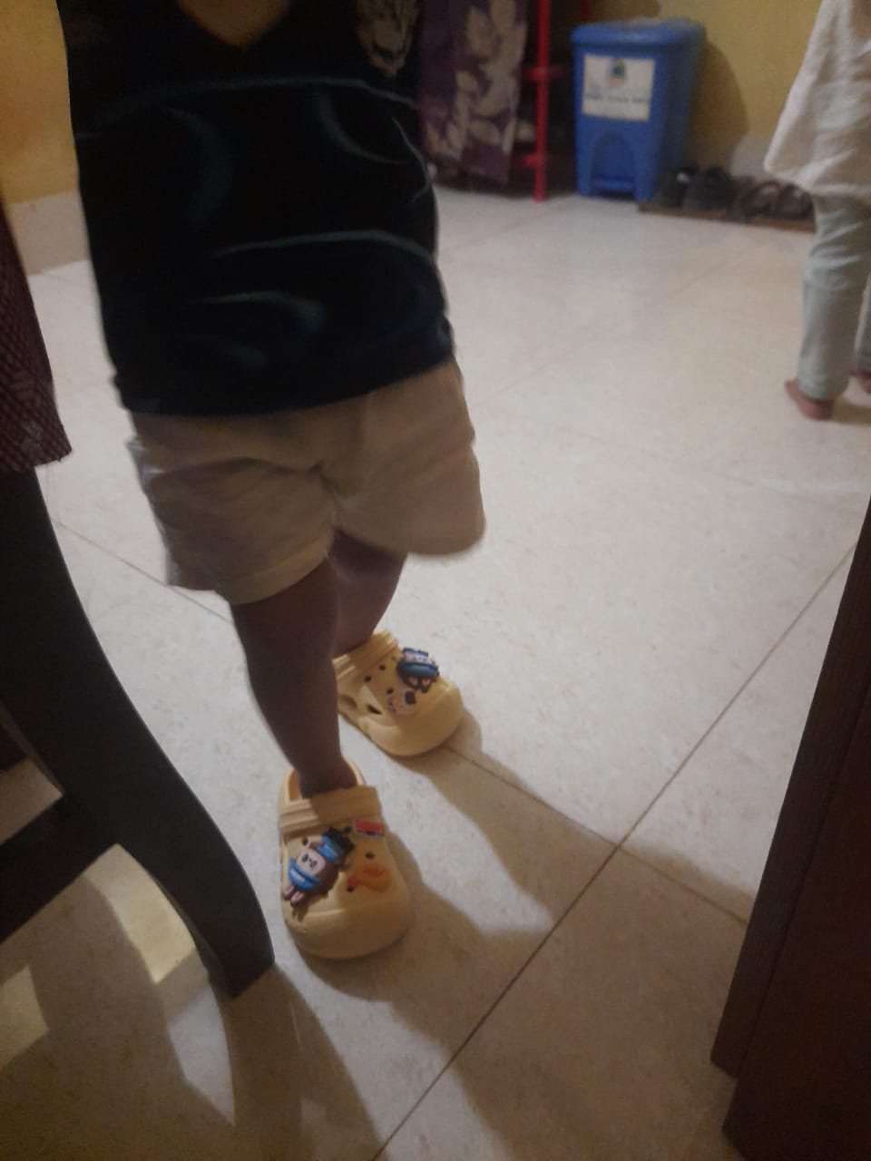 Toddler walking on a tiled floor in cream clog-style shoes with cartoon charms