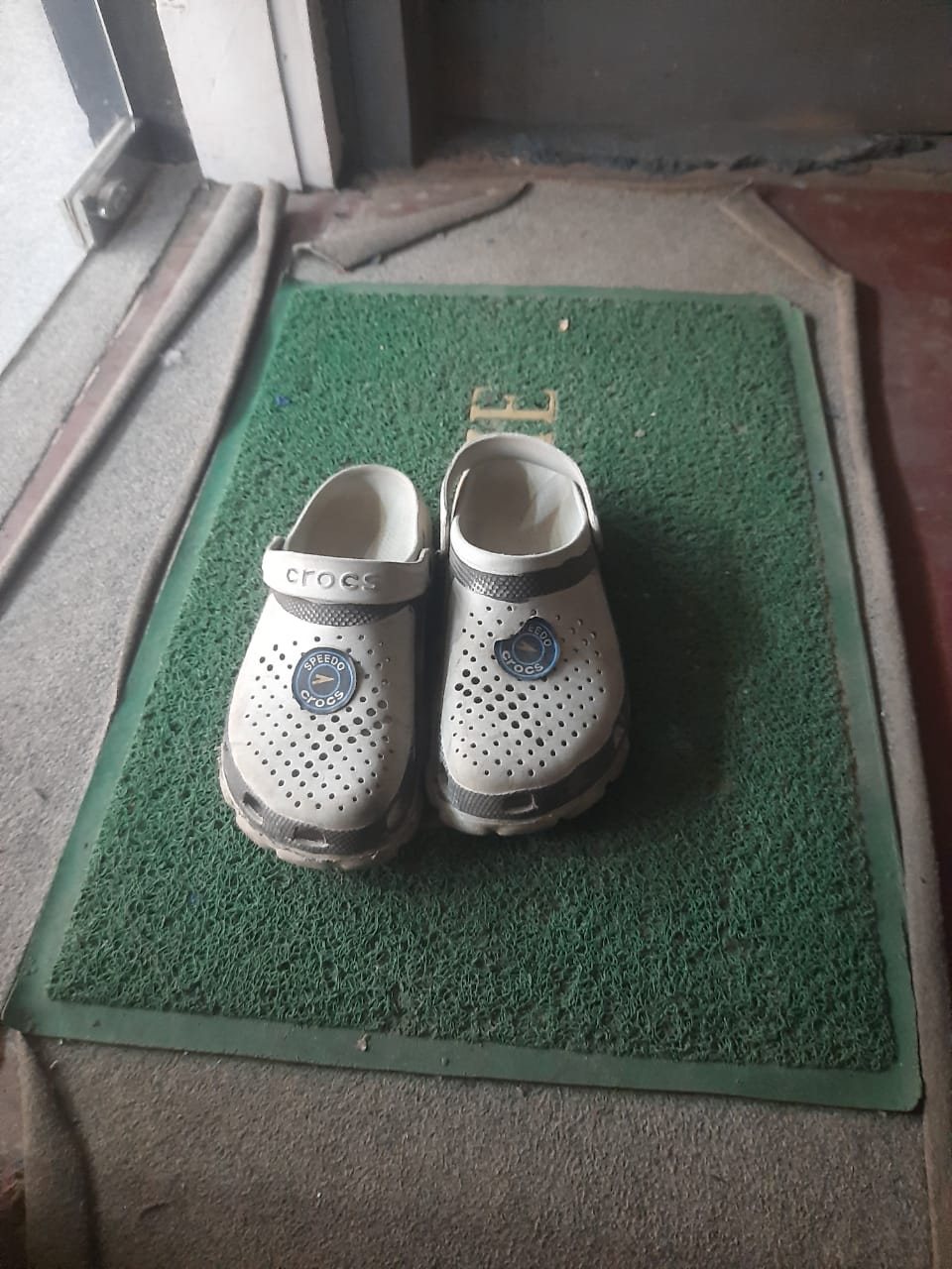 Pair of white and grey Crocs with round charms on a welcome mat after a day out
